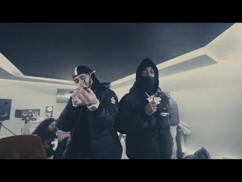 Two men stand in an indoor space, one wears a full face covering. A woman sits in the background. The scene has a grey color grading.