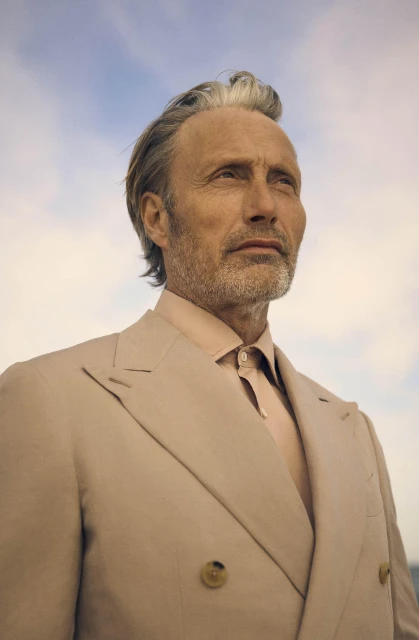 Portrait of middle-aged man with grey hair wearing a beige suit against a blue sky background, looking upward with a thoughtful expression.