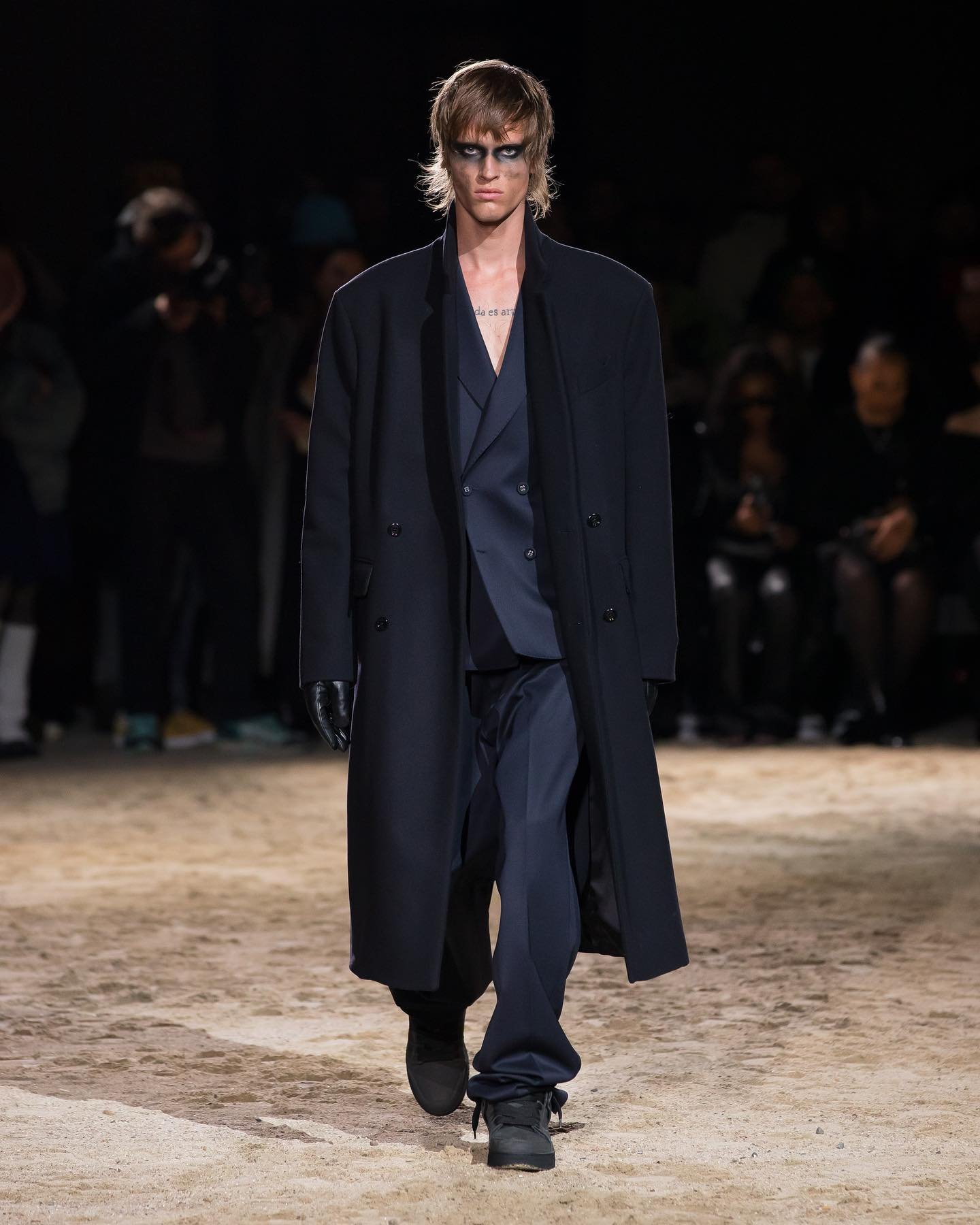 Male fashion model walks runway in black suit, coat, and black eye makeup, showcasing fashion design, runway style, and menswear trends.