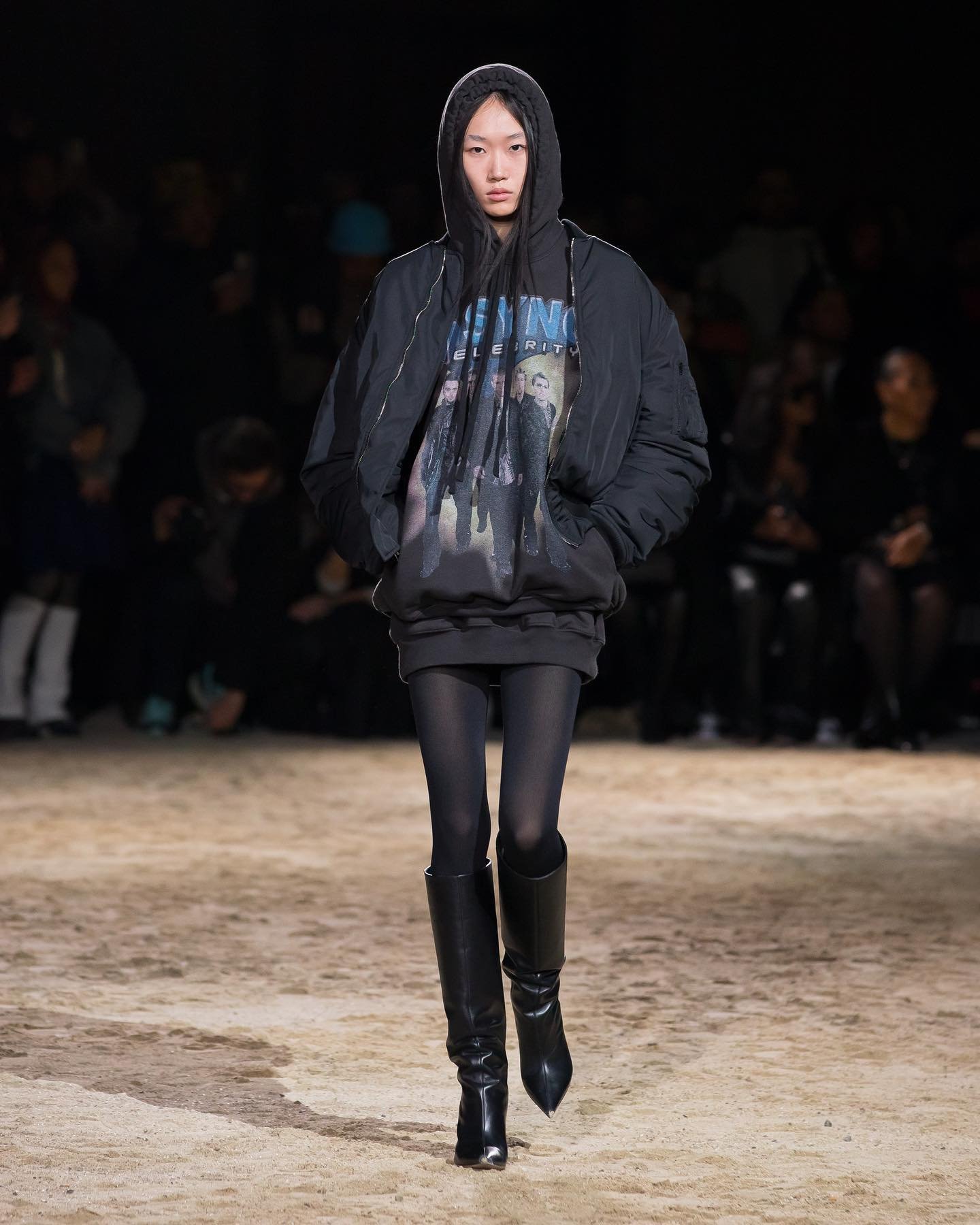 Runway model walks the catwalk in a black hooded sweatshirt, black puffer jacket, black tights, and black knee-high boots.