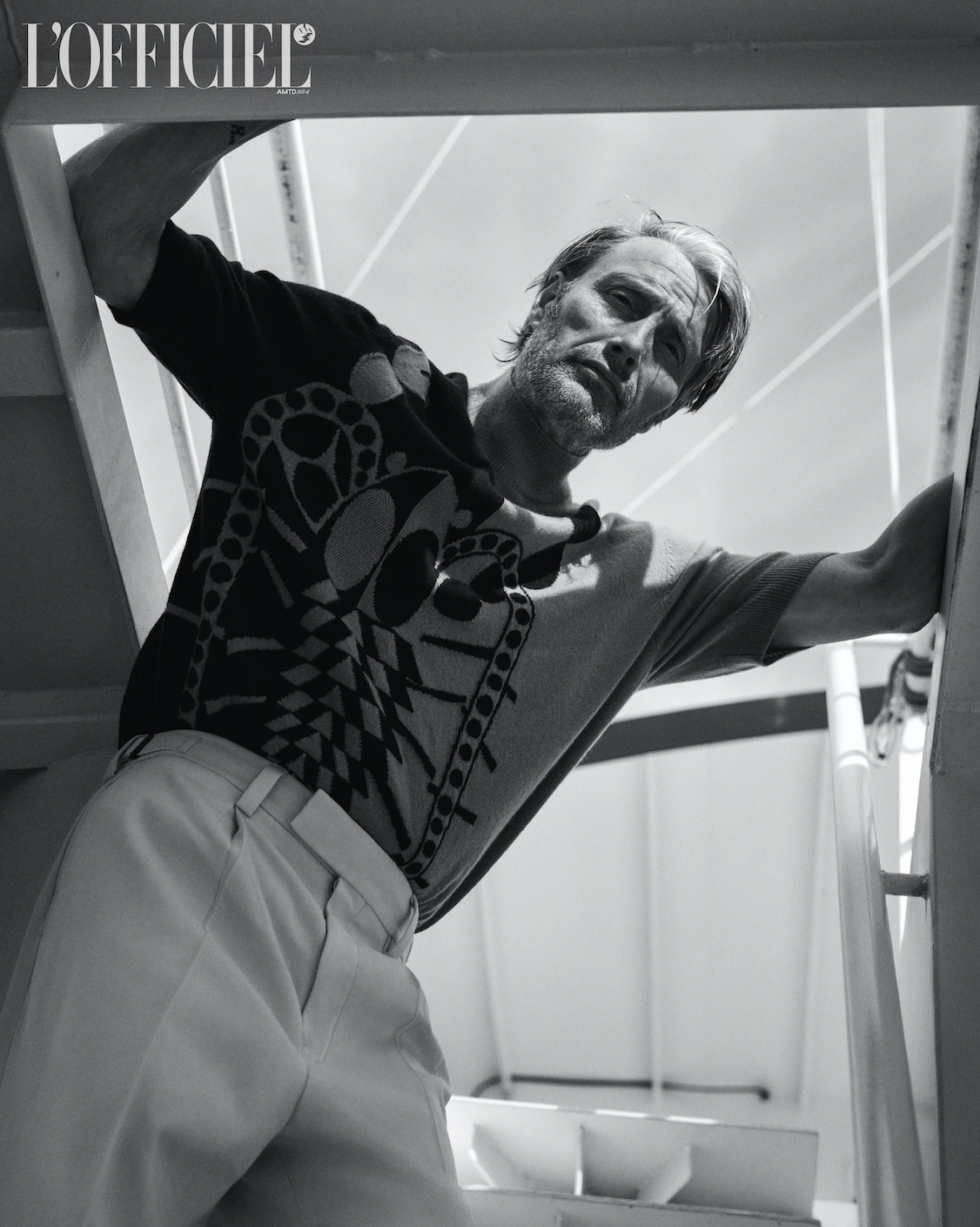 Black and white portrait of a middle-aged male model, leaning against a stair rail, wearing a patterned sweater and light trousers.
