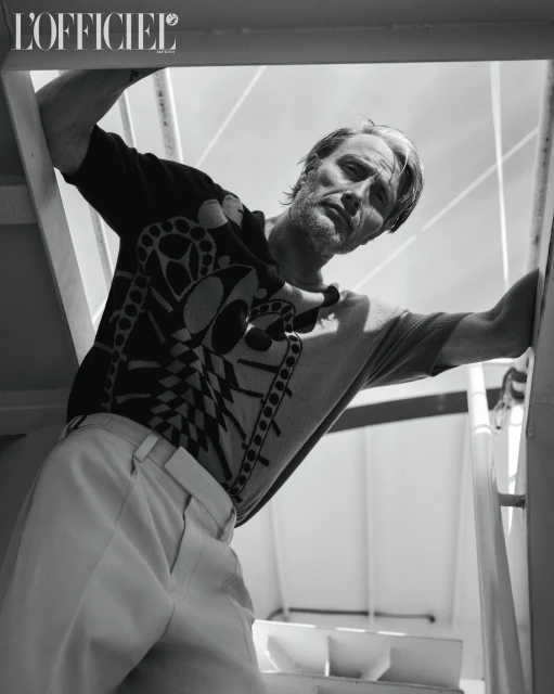 Black and white portrait of a middle-aged male model, leaning against a stair rail, wearing a patterned sweater and light trousers. 