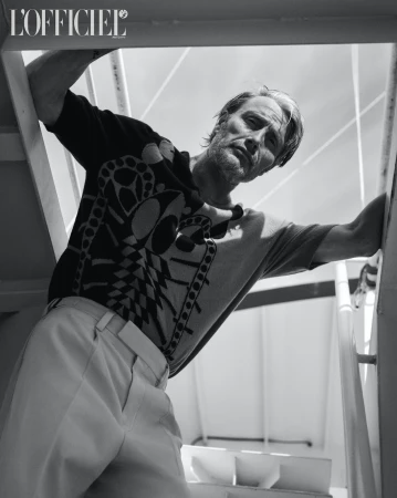 Black and white portrait of a middle-aged male model, leaning against a stair rail, wearing a patterned sweater and light trousers. 