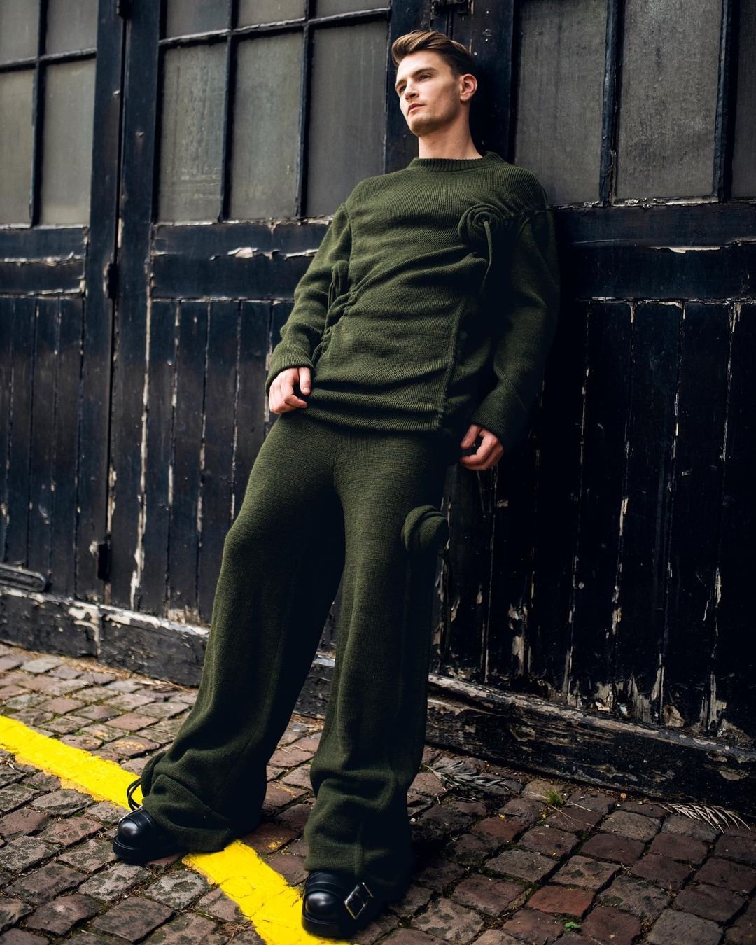Man in dark green knitwear poses against a dark industrial doorway, showcasing fashion and style in an outdoor setting with cobblestone.