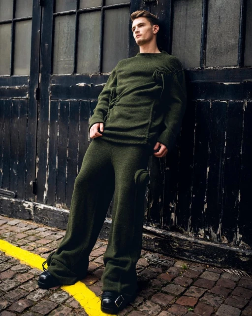 Man in dark green knitwear poses against a dark industrial doorway, showcasing fashion and style in an outdoor setting with cobblestone.