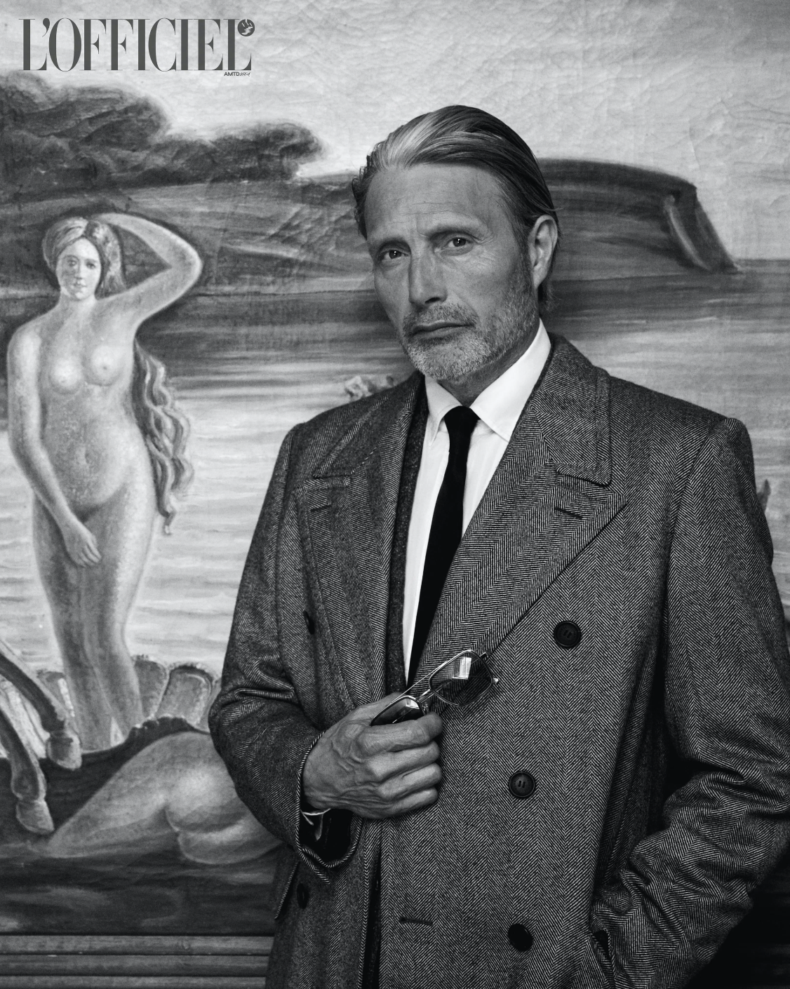 Monochrome portrait of Mads Mikkelsen wearing a double-breasted overcoat and holding sunglasses, posed in front of a classical painting.