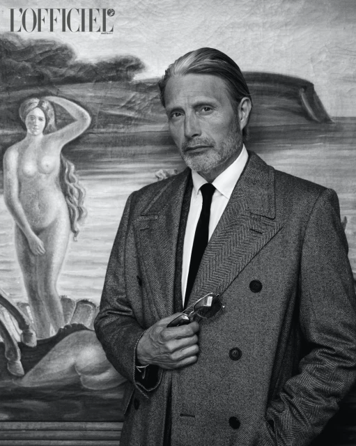 Monochrome portrait of Mads Mikkelsen wearing a double-breasted overcoat and holding sunglasses, posed in front of a classical painting.