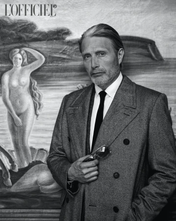 Monochrome portrait of Mads Mikkelsen wearing a double-breasted overcoat and holding sunglasses, posed in front of a classical painting.