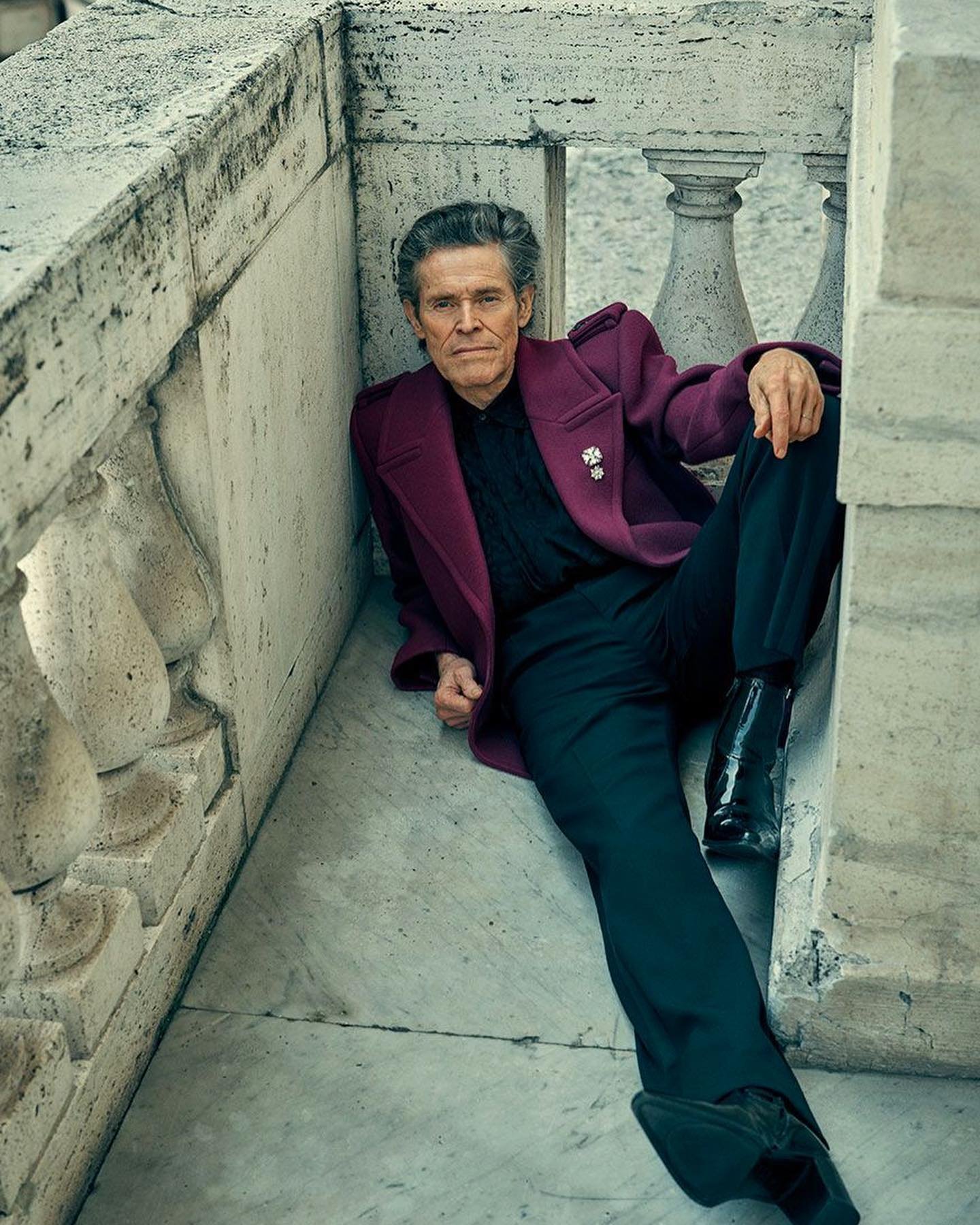 Willem Dafoe, in a maroon coat and dark pants, poses on a stone ledge with balustrades, with a relaxed expression.