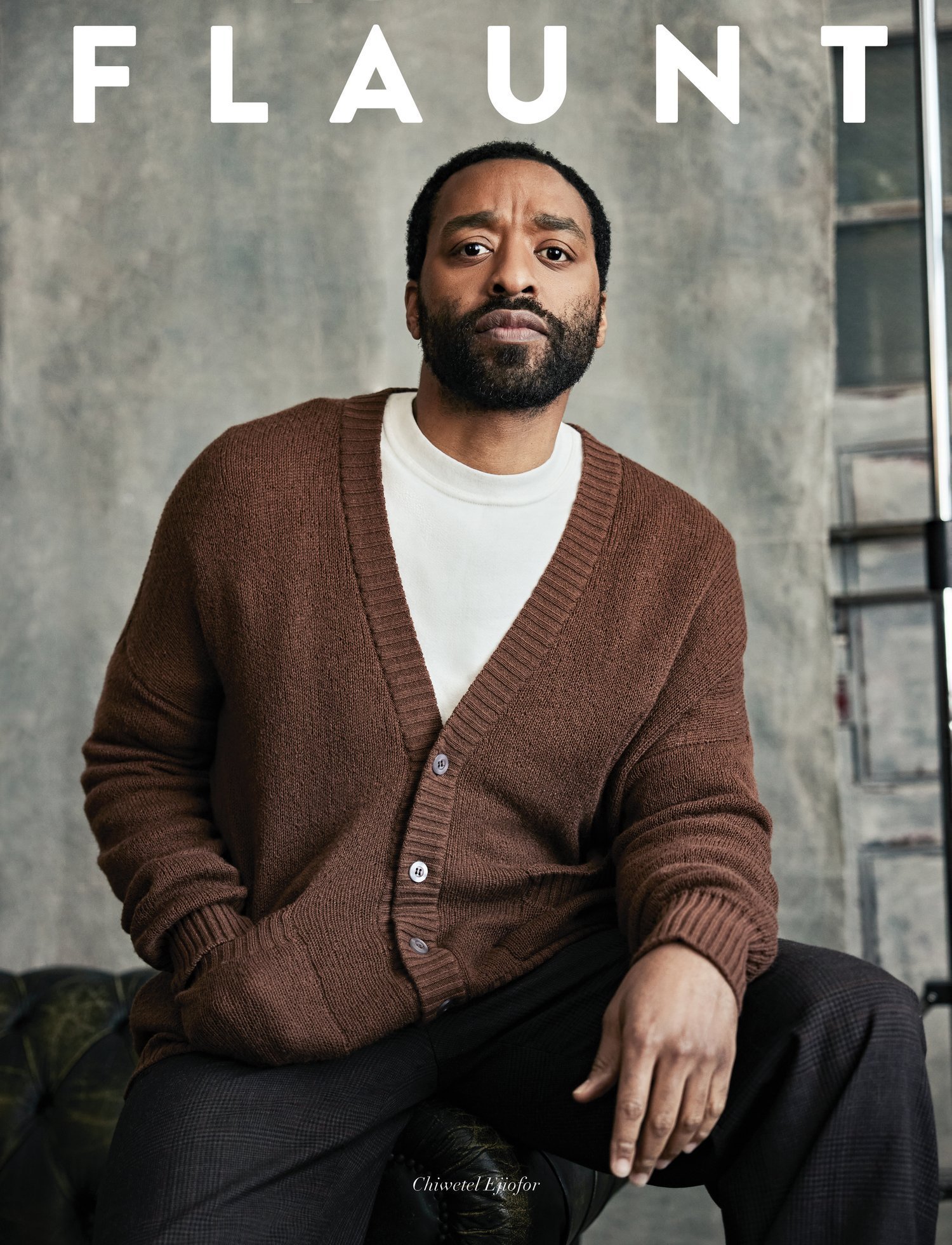 Portrait of adult male wearing a brown cardigan sweater and white shirt, sitting on a green leather seat in a neutral indoor space.
