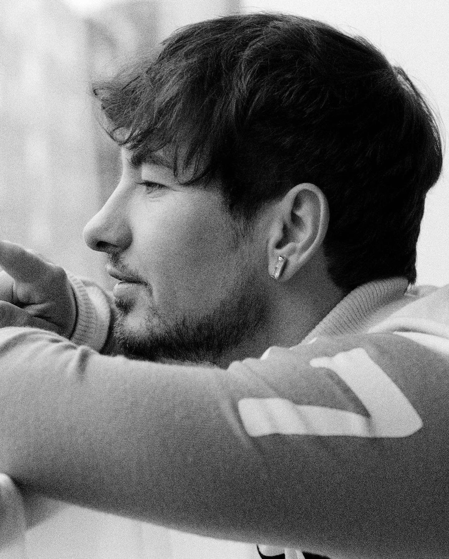 Monochrome portrait of a young adult male looking away with his arm resting on a surface, showcasing his dark hair and earring.