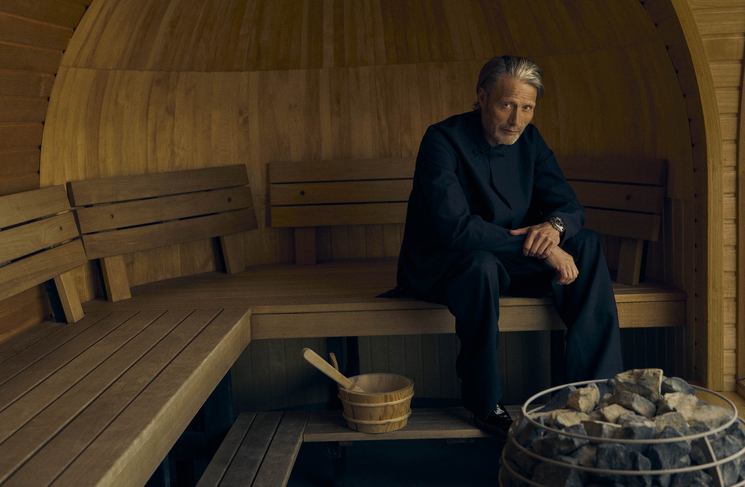 Portrait of Mads Mikkelsen in a sauna, seated on a wooden bench with a bucket and ladle nearby, with a wood-paneled interior.