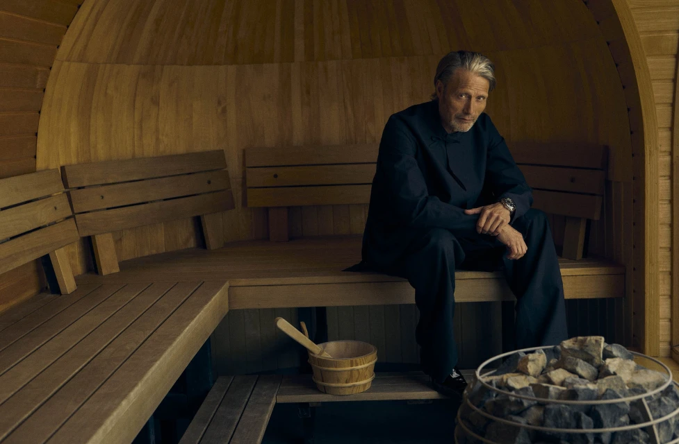 Portrait of Mads Mikkelsen in a sauna, seated on a wooden bench with a bucket and ladle nearby, with a wood-paneled interior.