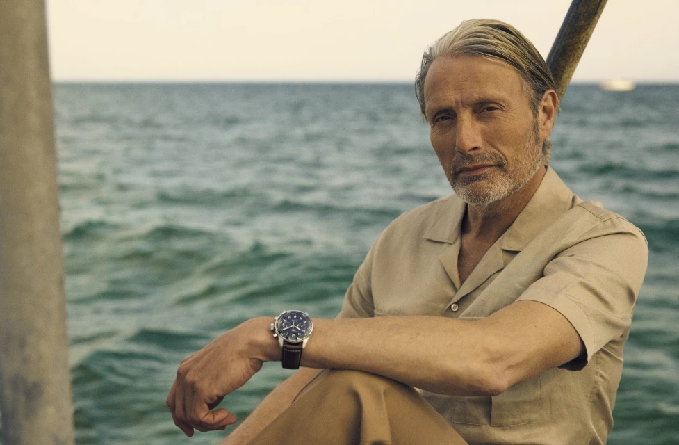 Mature man with short grey hair sits outdoors by the ocean wearing a short-sleeved shirt and brown pants, with a watch on his wrist.