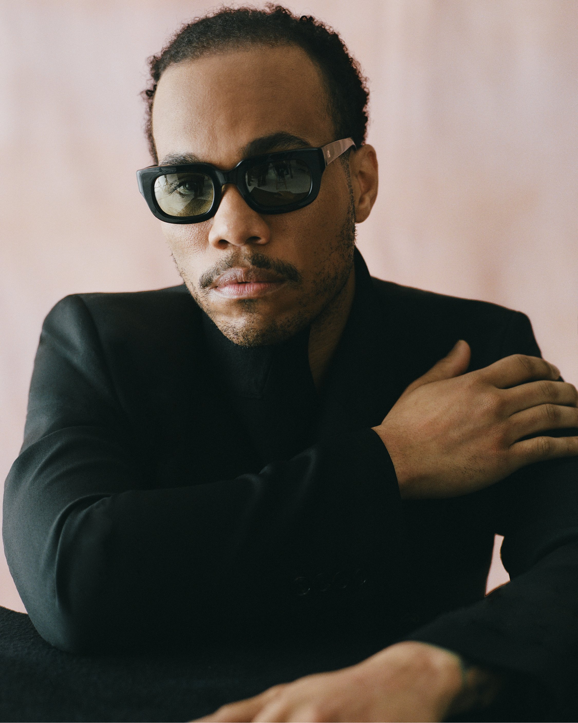 Portrait of a stylish black man with sunglasses, wearing a black suit against a pastel pink background in a studio setting.