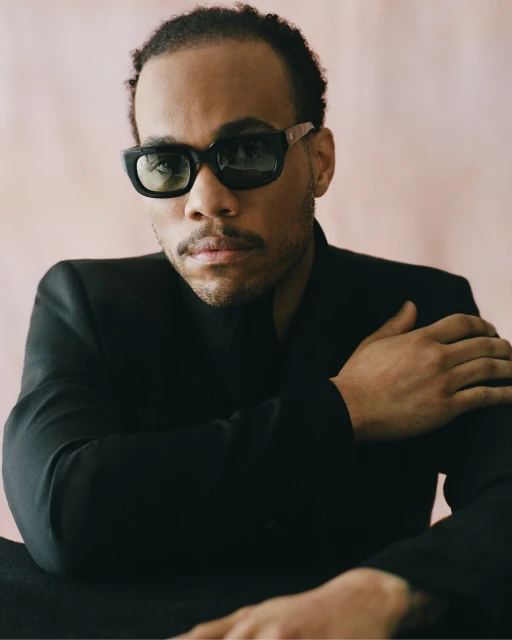 Portrait of a stylish black man with sunglasses, wearing a black suit against a pastel pink background in a studio setting.