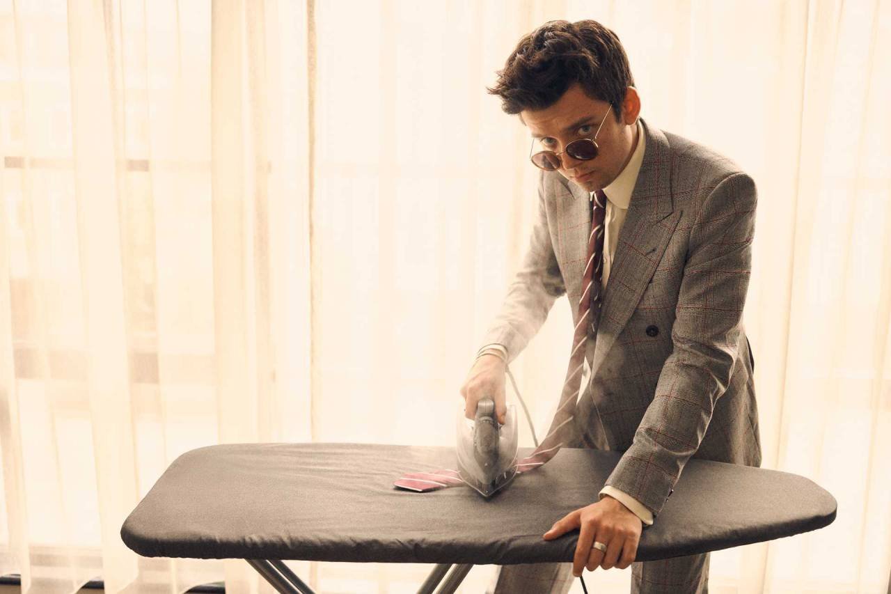 Adult male in a grey suit and sunglasses irons a tie on a black ironing board against a blurred background of curtains.