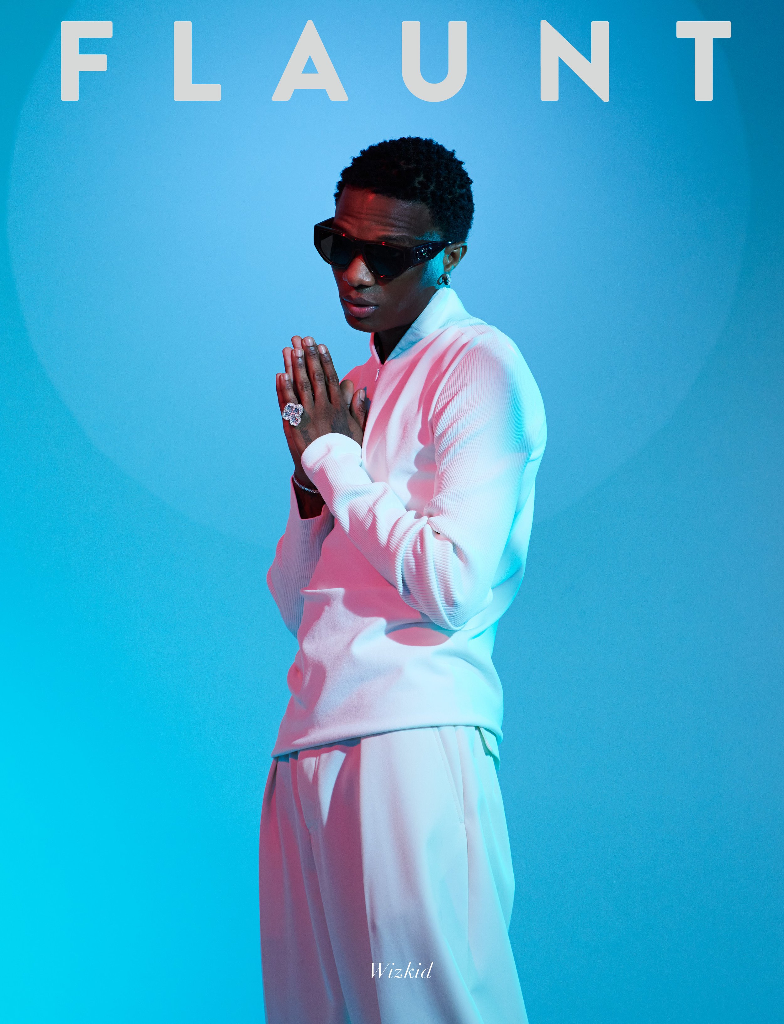 Man with sunglasses and a ring posing in front of a blue background with the word FLAUNT in the upper left corner.