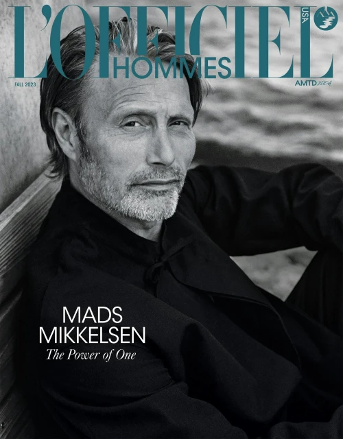 Mads Mikkelsen wearing a black shirt with styled hair and stubble against a neutral textured background for L'Officiel Hommes.