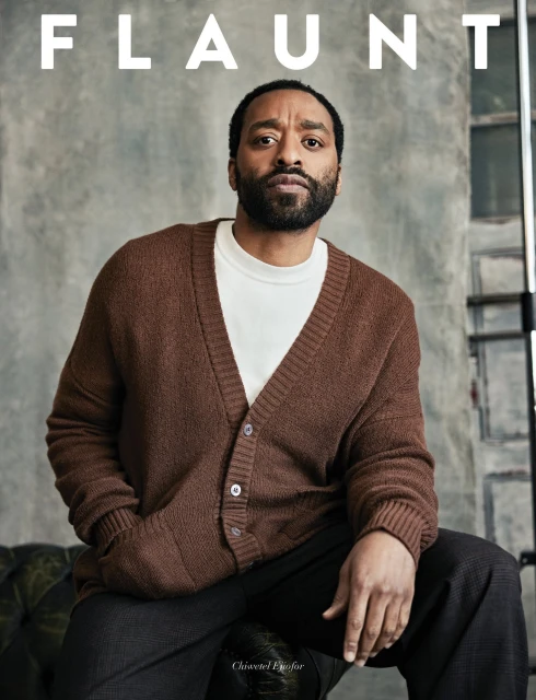 Chiwetel Ejiofor wearing a brown knit cardigan and dark trousers against a textured grey backdrop for Flaunt magazine