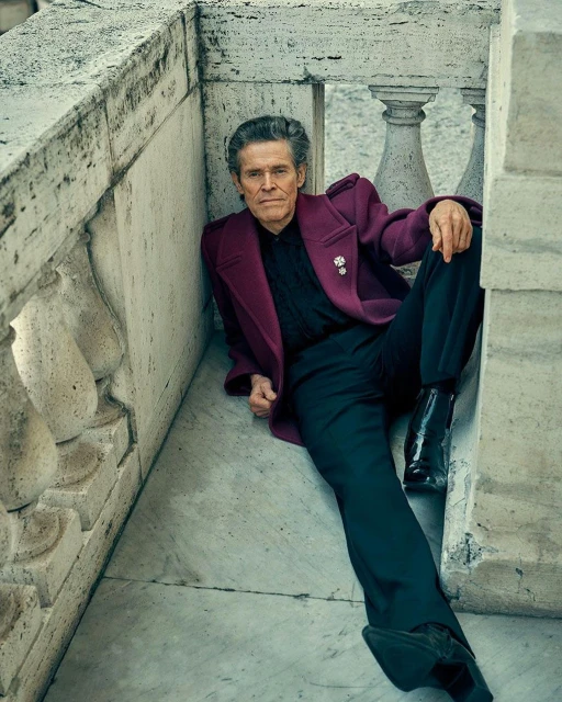 Willem Dafoe wearing a magenta jacket and black trousers seated on a stone balcony, styled by Jay Hines