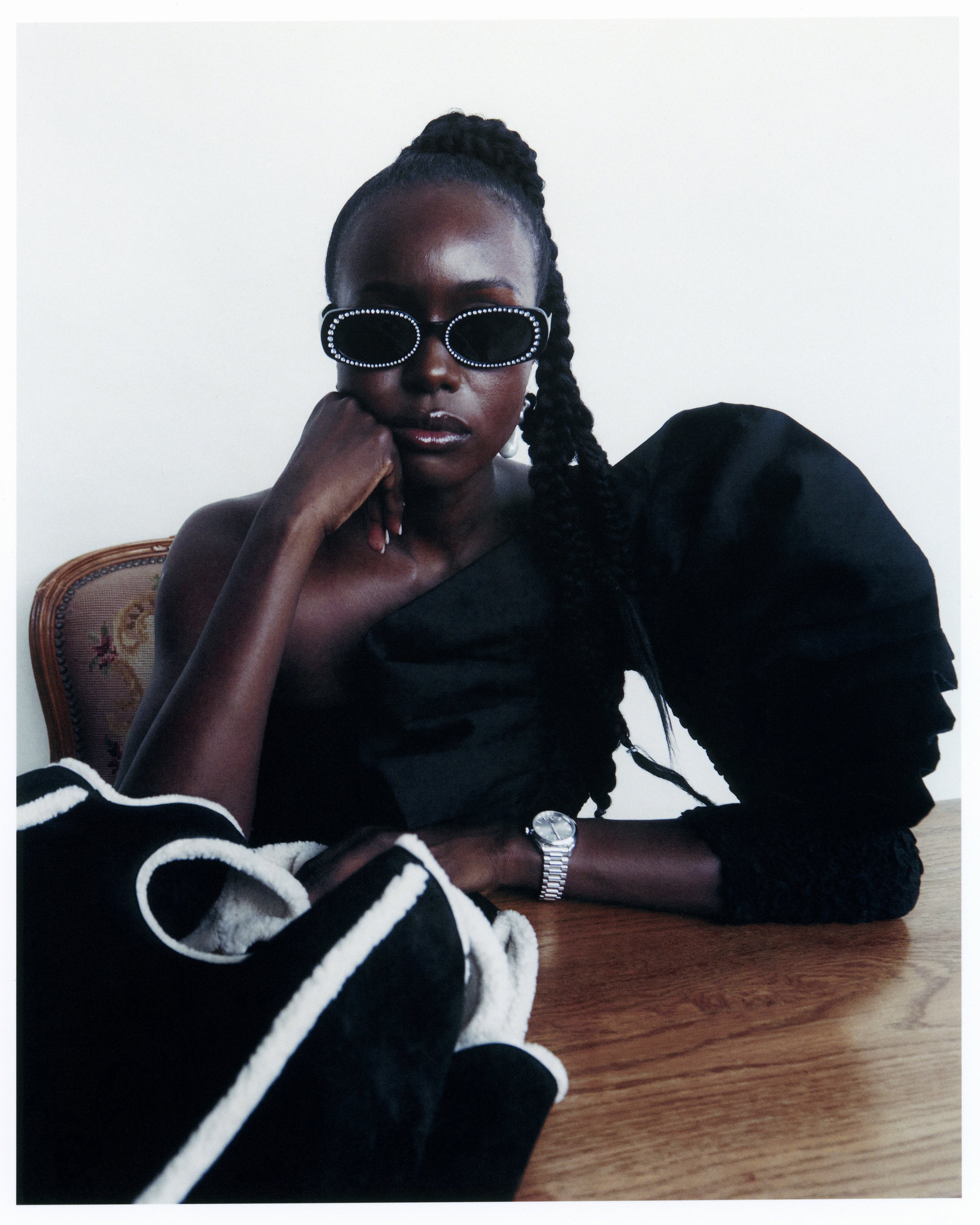 Portrait of an African woman in a black dress with puffed sleeve at a wooden table, wearing jeweled sunglasses in a studio setting.