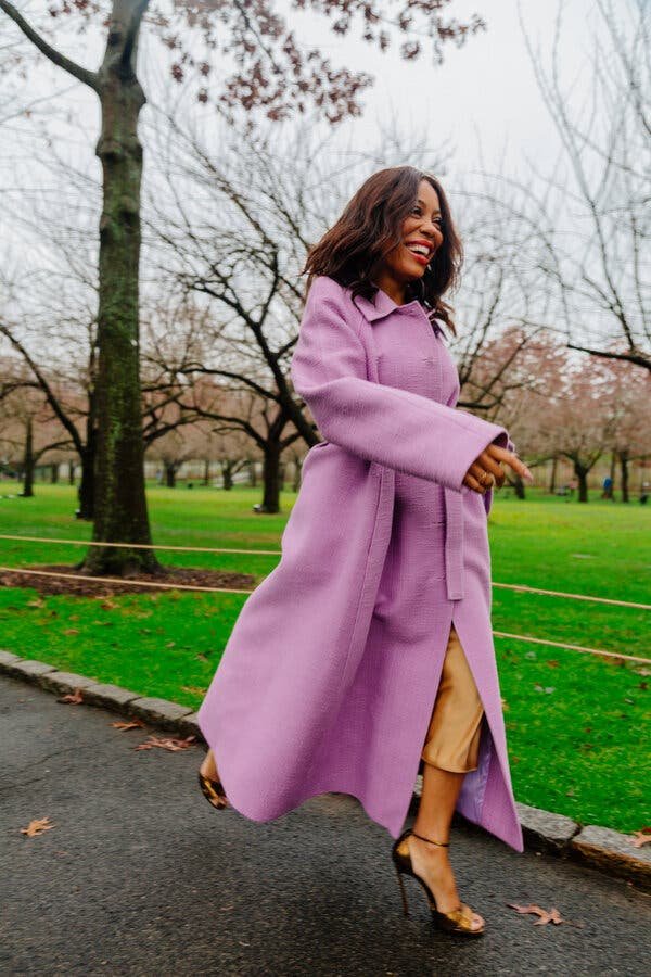 Stylish woman in a long lilac coat walks along a park path, smiling, with trees and green grass in the background.