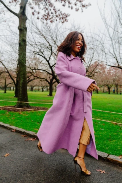 Stylish woman in a long lilac coat walks along a park path, smiling, with trees and green grass in the background.