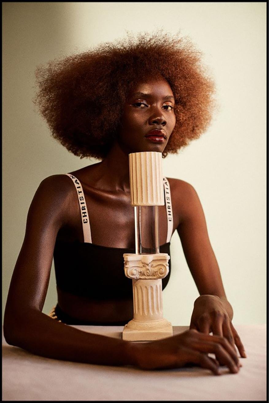 Portrait of Black fashion model posing indoors with clear vase and neoclassical pedestal, wearing black top and brown afro hairstyle.