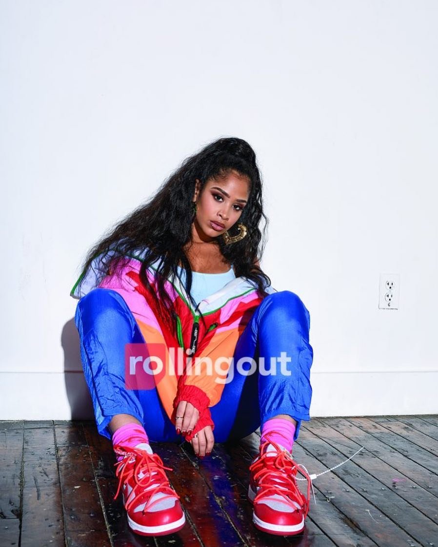 A woman with long, dark hair, wearing a colorful jacket and blue pants, sits on a wood floor in front of a white wall, smiling slightly.