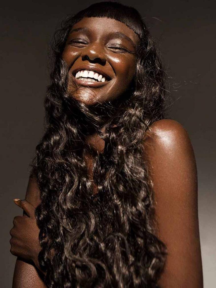 Close-up portrait of a smiling African-American woman with curly dark hair, shiny skin, and a toothy smile against a dark background.