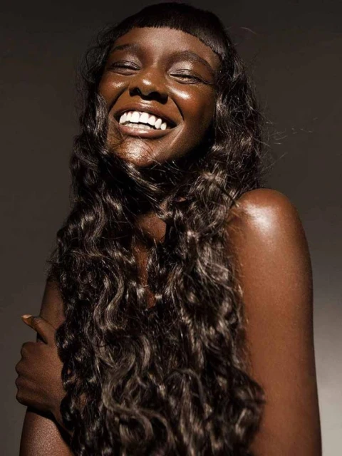 Close-up portrait of a smiling African-American woman with curly dark hair, shiny skin, and a toothy smile against a dark background.