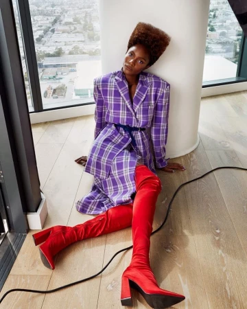 Woman in plaid blazer and red boots sitting against a column, with city views visible through a window in an indoor setting.