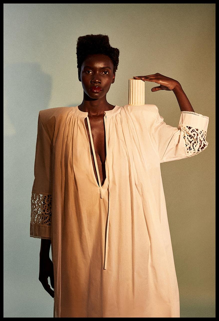Striking image of a stylish dark-skinned woman in a flowing dress, posing with a textured architectural column in a fashion editorial.