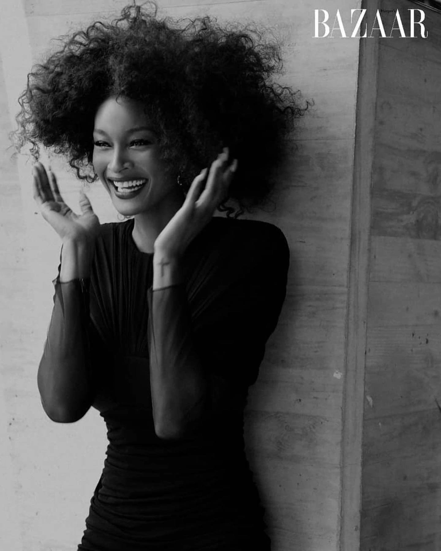 Black and white portrait of a smiling Black woman with afro hairstyle wearing a black dress in front of a textured wall.