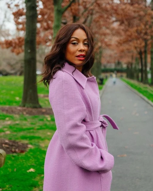 Outdoor portrait of a woman in a lilac coat standing on a path lined with trees in autumn, featuring fallen leaves and soft lighting.