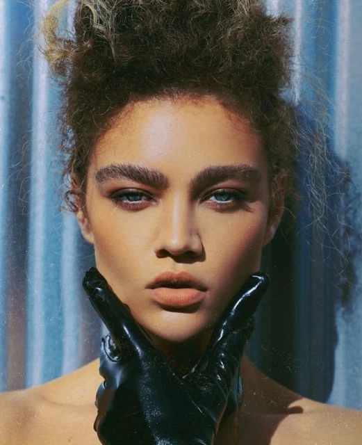 Close-up portrait of an adult female with curly hair and blue eyes, wearing black gloves, against a textured blue background.