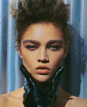 Close-up portrait of an adult female with curly hair and blue eyes, wearing black gloves, against a textured blue background.