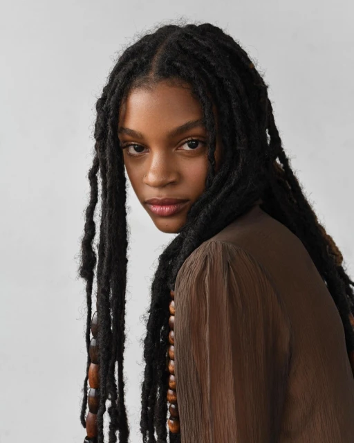 Portrait of an African American woman with dreadlocks, wearing a brown top and beaded necklace, gazing at the camera against a grey backdrop.