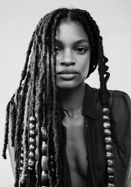 Portrait of a Black woman with locs and beads in a semi-sheer dark top, showcasing a close-up with a neutral expression against a light background.