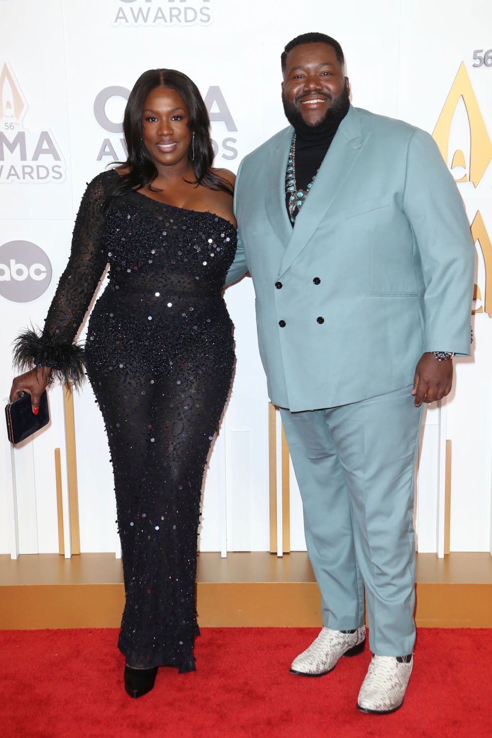 A formal full shot of a man and woman posing on a red carpet, dressed in formal attire, under bright lighting, with a backdrop of awards signage.