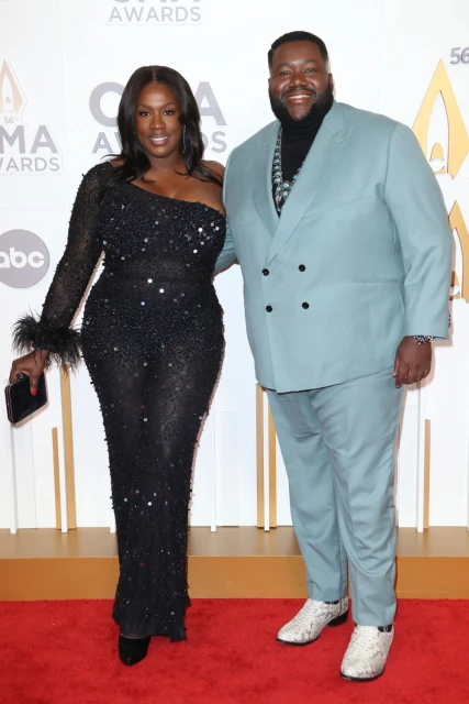 A formal full shot of a man and woman posing on a red carpet, dressed in formal attire, under bright lighting, with a backdrop of awards signage.