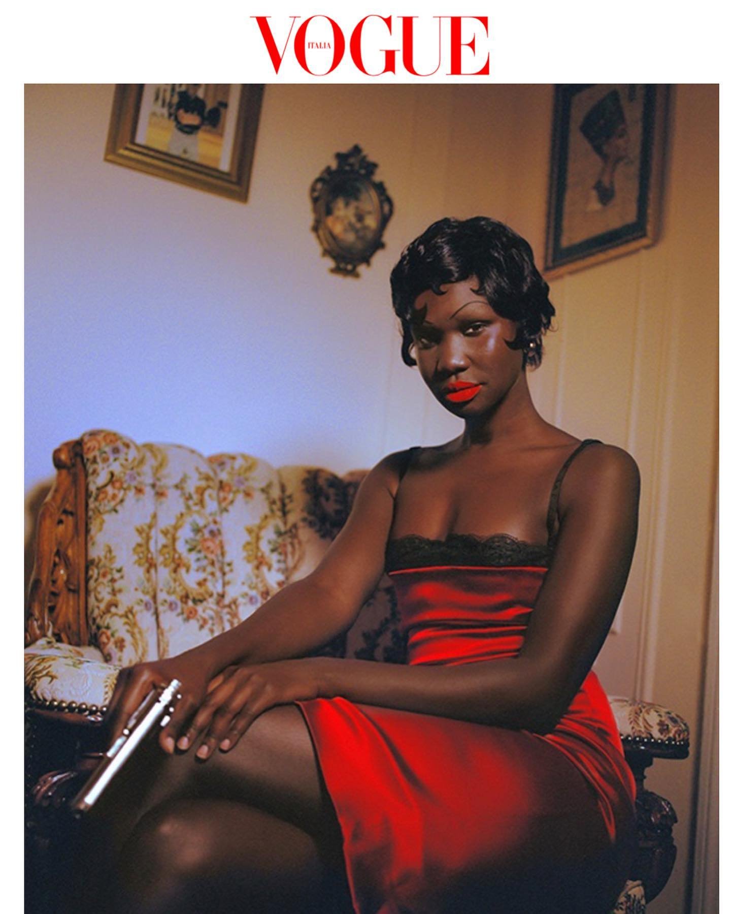 Glamorous African-American woman in red dress poses with handgun on floral settee for Vogue cover; framed art and logo visible.