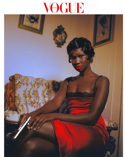 Glamorous African-American woman in red dress poses with handgun on floral settee for Vogue cover; framed art and logo visible.