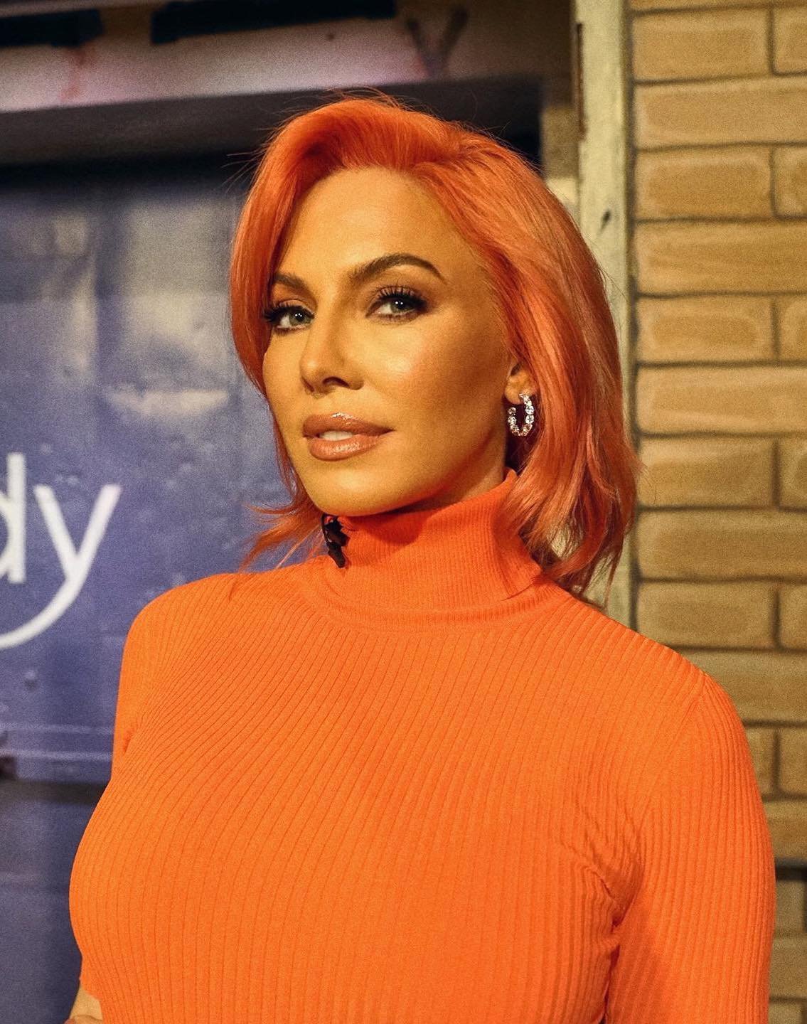 Close-up shot of an adult woman with orange hair wearing a bright orange turtleneck sweater in front of a brick wall.