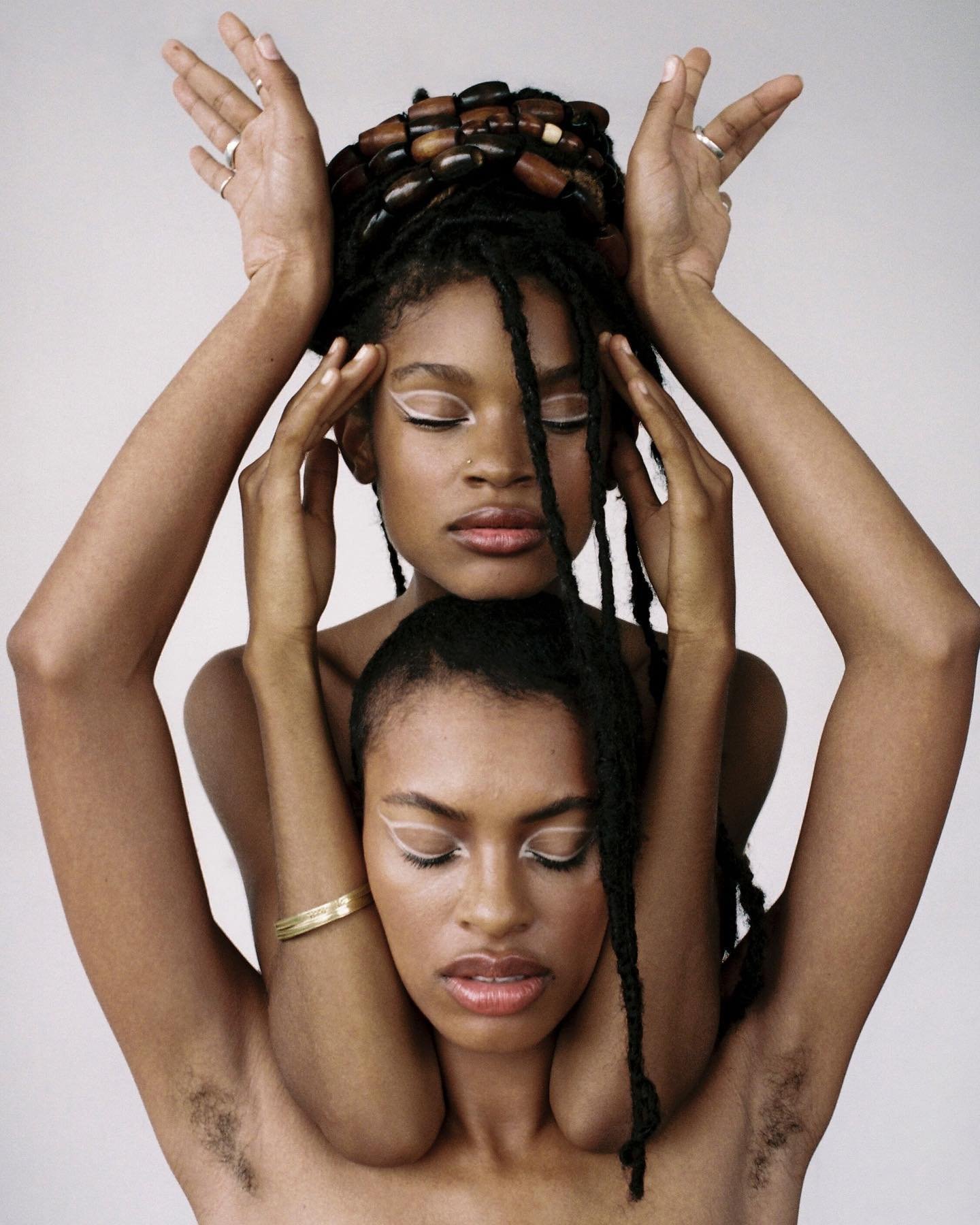 Two Black women in a portrait. The woman on the bottom has her eyes closed and a gold bangle while the woman on top has dreadlocks.