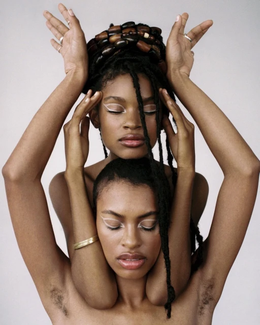 Two Black women in a portrait. The woman on the bottom has her eyes closed and a gold bangle while the woman on top has dreadlocks.