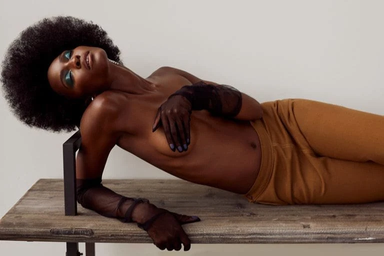 Dark-skinned woman with an afro, wearing sheer gloves and pants, is reclining on a wooden surface in a fashion or beauty editorial photo.