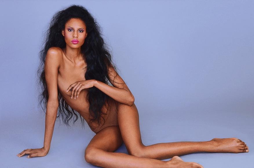 Woman with long dark hair poses in a nude body suit against a light blue backdrop in a beauty and fashion style photograph.
