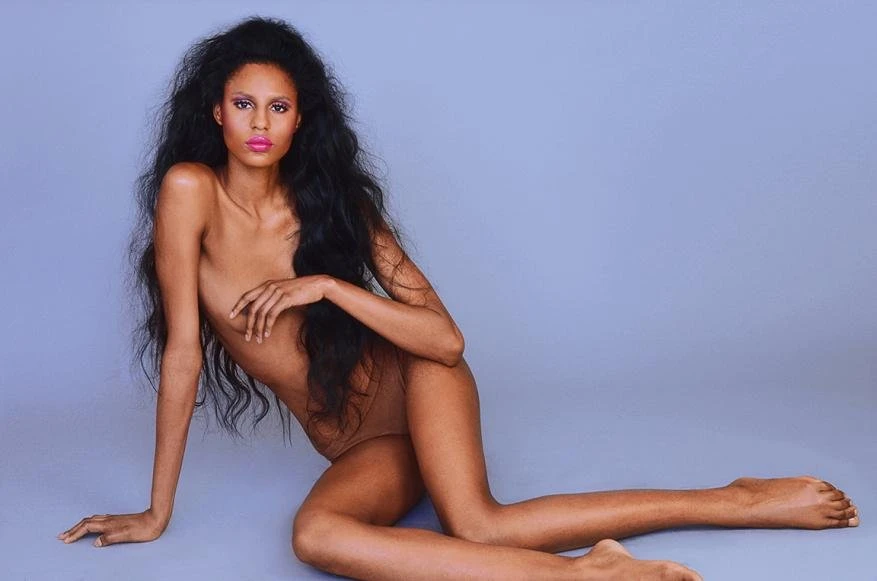 Woman with long dark hair poses in a nude body suit against a light blue backdrop in a beauty and fashion style photograph.